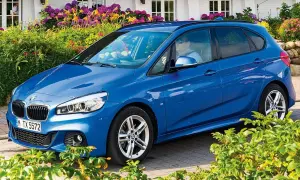 Blauer BMW 218i Active Tourer vor blühenden Sträuchern.