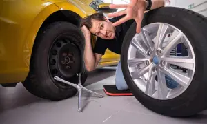 Mann mit Reifen und Werkzeug, der frustriert aussieht. Die **Handbremse beim Reifenwechsel** ist angezogen, um ein Wegrollen zu verhindern.