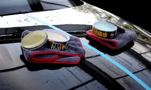 Zwei Dosen Autowachs und Mikrofasertücher liegen auf einer glänzenden Motorhaube. Ein blauer Streifen markiert den Bereich, der für die Autopflege vorbereitet wird.