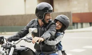 Ein Mann mit Bart und schwarzer Lederjacke nimmt ein Kind auf seinem Motorrad mit. Beide tragen Helme und lächeln sich an.