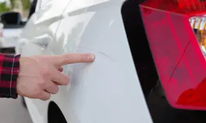 Finger zeigt auf Kratzer am weißen Auto, vermutlich verursacht durch die Waschanlage.