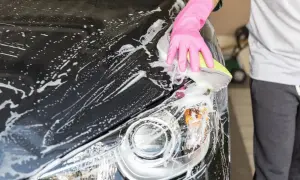 Eine Hand in pinker Handschuh-Hülle reinigt mit einem Schwamm das nasse, schwarze Auto. Schaum bedeckt die Motorhaube und den Scheinwerfer.