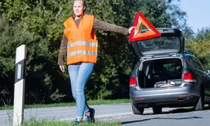 Frau mit Warnweste und Warndreieck neben offenem Kofferraum. Sie bereitet sich auf das auto aufbocken vor, um das Fahrzeug zu reparieren.