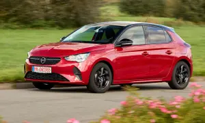 Roter Opel Corsa F fährt auf einer Straße, umgeben von grünen Feldern und blühenden Blumen. Keine Probleme mit diesem flotten Flitzer!
