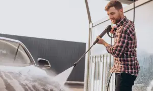 Ein Mann mit kariertem Hemd nutzt einen Hochdruckreiniger, um sein Auto zu waschen.