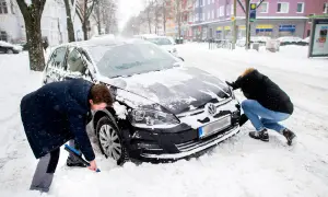 Zwei Personen schaufeln Schnee vom auto im winter. Ein Mann mit blauer Bürste reinigt das Rad, eine Frau schiebt die Motorhaube.