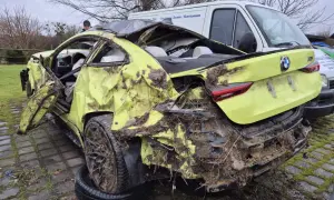 Ein gelber BMW mit Totalschaden, stark beschädigt und mit Schlamm bedeckt, steht neben einem weißen Lieferwagen.