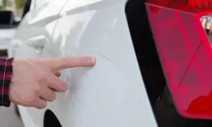 Finger zeigt auf tiefe Kratzer bis auf Grundierung am weißen Auto.