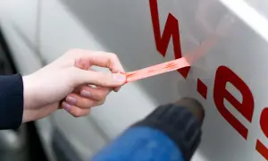 Hand zieht roten Aufkleber vom Auto ab, während ein Heißluftföhn das Entfernen erleichtert.