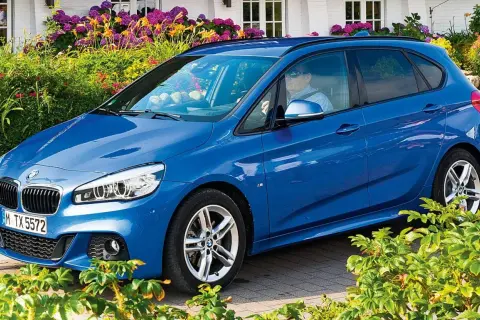 Blauer BMW 218i Active Tourer vor blühenden Sträuchern.