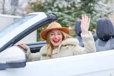 Fröhliche Frau winkt aus einem weißen Cabrio im Winter, während Schneeflocken fallen.