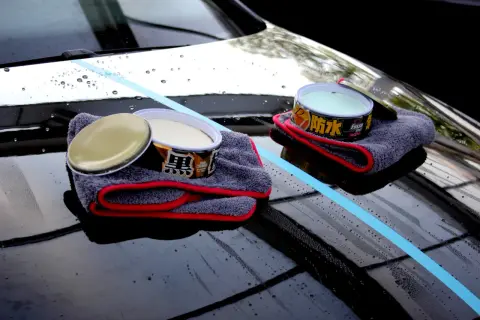 Zwei Dosen Autowachs und Mikrofasertücher liegen auf einer glänzenden Motorhaube. Ein blauer Streifen markiert den Bereich, der für die Autopflege vorbereitet wird.