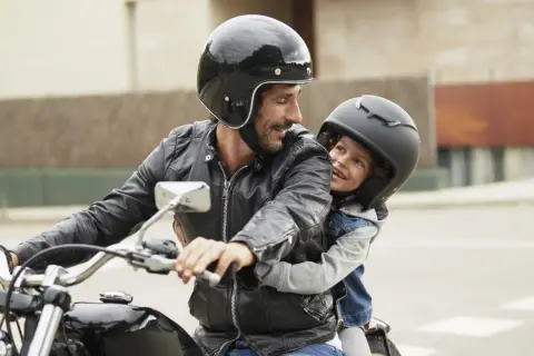 Ein Mann mit Bart und schwarzer Lederjacke nimmt ein Kind auf seinem Motorrad mit. Beide tragen Helme und lächeln sich an.