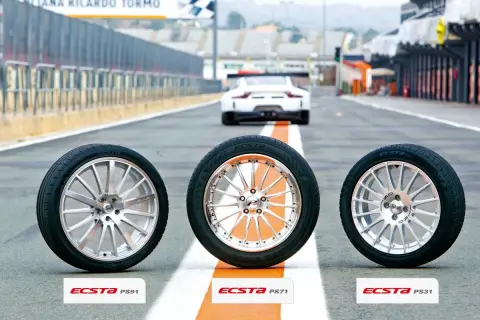 Drei Kumho Ecsta PS71 Reifen stehen auf einer Rennstrecke, bereit für einen Test. Ein weißer Sportwagen fährt im Hintergrund.
