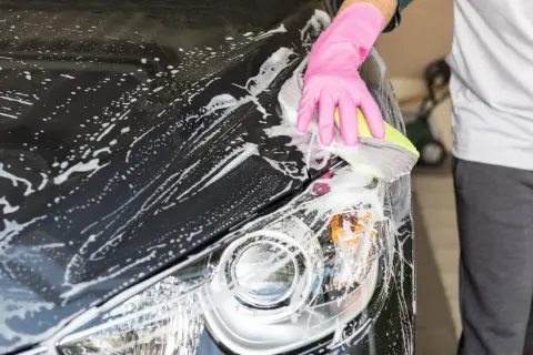 Eine Hand in pinker Handschuh-Hülle reinigt mit einem Schwamm das nasse, schwarze Auto. Schaum bedeckt die Motorhaube und den Scheinwerfer.