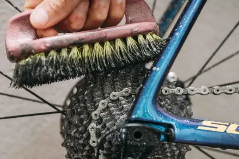Mit Hausmitteln die Fahrradkette reinigen: Eine Bürste schrubbt die Kette und Kassette des glänzenden blauen Fahrradrahmens.