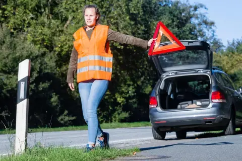 Frau mit Warnweste und Warndreieck neben offenem Kofferraum. Sie bereitet sich auf das auto aufbocken vor, um das Fahrzeug zu reparieren.
