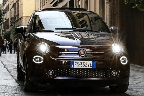 Dunkelbrauner Fiat 500 mit leuchtenden Scheinwerfern auf einer Kopfsteinpflasterstraße. Ein paar Leute gehen im Hintergrund vorbei. Keine sichtbaren Fiat 500 Probleme.