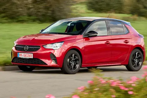 Roter Opel Corsa F fährt auf einer Straße, umgeben von grünen Feldern und blühenden Blumen. Keine Probleme mit diesem flotten Flitzer!
