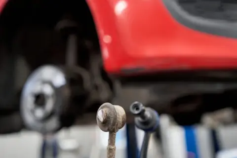 Zwei Koppelstangen liegen vor einem roten Auto. Eine Koppelstange ist defekt und muss ersetzt werden.