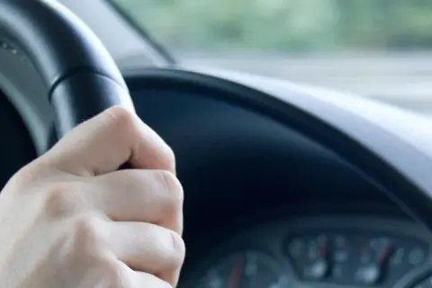 Hand am Lenkrad, das leicht flattert. Die Straße zieht vorbei, ein Gefühl von Bewegung.