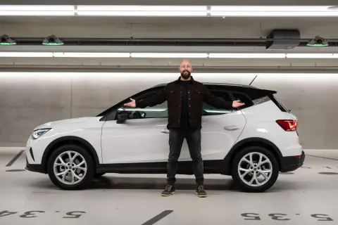 Mann neben weißem Seat Arona in Tiefgarage, lächelnd mit ausgestreckten Armen. Keine Seat Arona Probleme hier.