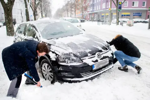 Zwei Personen schaufeln Schnee vom auto im winter. Ein Mann mit blauer Bürste reinigt das Rad, eine Frau schiebt die Motorhaube.