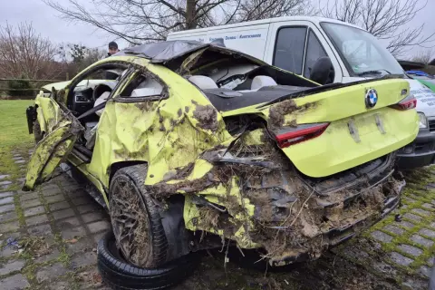 Ein gelber BMW mit Totalschaden, stark beschädigt und mit Schlamm bedeckt, steht neben einem weißen Lieferwagen.