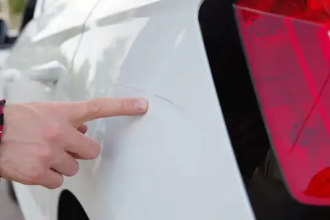 Finger zeigt auf tiefe Kratzer bis auf Grundierung am weißen Auto.