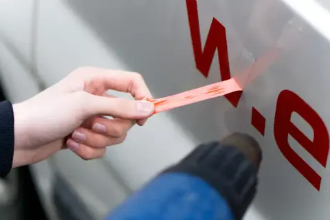 Hand zieht roten Aufkleber vom Auto ab, während ein Heißluftföhn das Entfernen erleichtert.