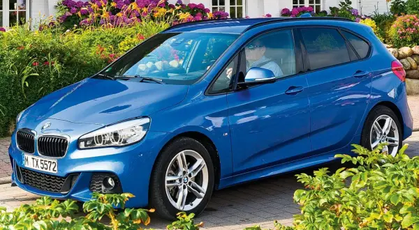 Blauer BMW 218i Active Tourer vor blühenden Sträuchern.