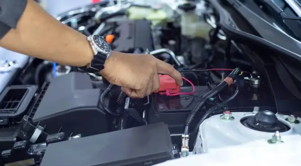 Hand mit Uhr lädt die Autobatterie ohne Abklemmen. Roter Stecker wird auf den Pluspol gesetzt.
