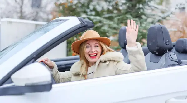 Fröhliche Frau winkt aus einem weißen Cabrio im Winter, während Schneeflocken fallen.