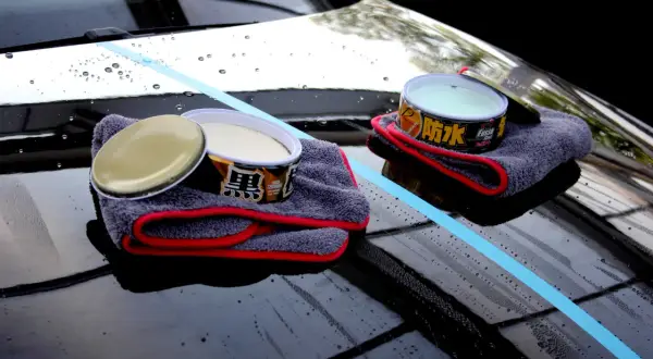 Zwei Dosen Autowachs und Mikrofasertücher liegen auf einer glänzenden Motorhaube. Ein blauer Streifen markiert den Bereich, der für die Autopflege vorbereitet wird.