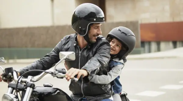 Ein Mann mit Bart und schwarzer Lederjacke nimmt ein Kind auf seinem Motorrad mit. Beide tragen Helme und lächeln sich an.