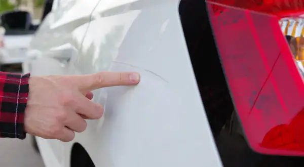 Finger zeigt auf Kratzer am weißen Auto, vermutlich verursacht durch die Waschanlage.