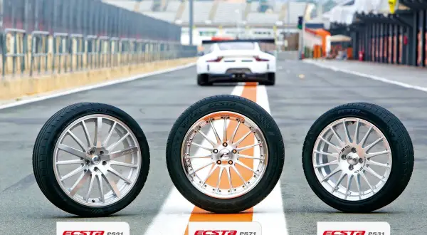 Drei Kumho Ecsta PS71 Reifen stehen auf einer Rennstrecke, bereit für einen Test. Ein weißer Sportwagen fährt im Hintergrund.