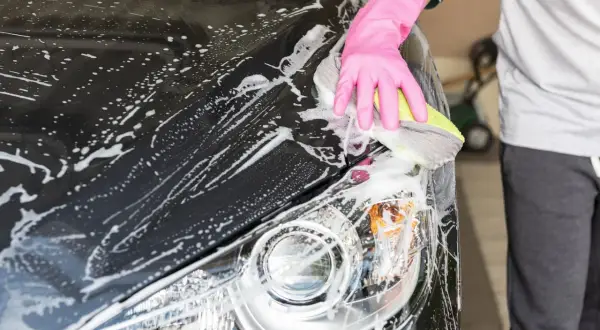 Eine Hand in pinker Handschuh-Hülle reinigt mit einem Schwamm das nasse, schwarze Auto. Schaum bedeckt die Motorhaube und den Scheinwerfer.
