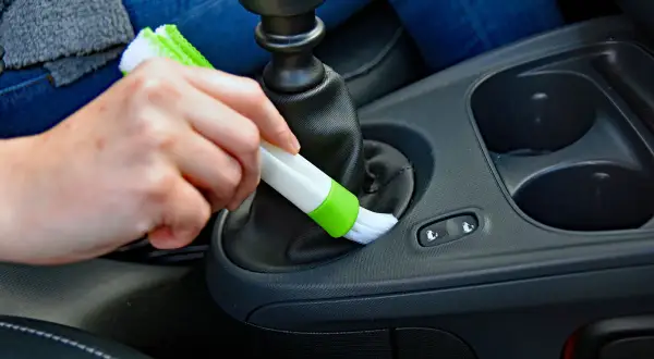 Hand reinigt mit Bürste den Bereich um den Schalthebel, um Staub im Auto zu entfernen.