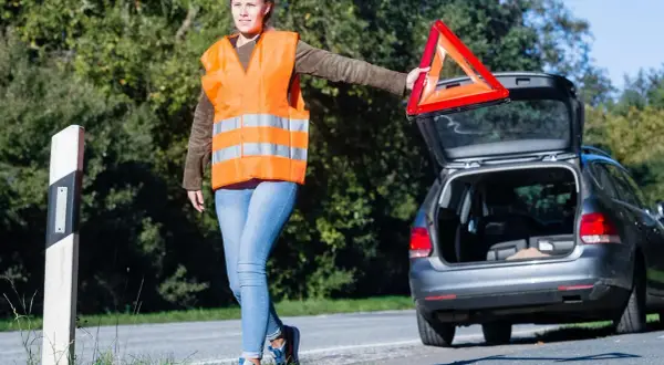 Frau mit Warnweste und Warndreieck neben offenem Kofferraum. Sie bereitet sich auf das auto aufbocken vor, um das Fahrzeug zu reparieren.