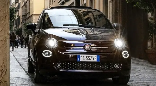 Dunkelbrauner Fiat 500 mit leuchtenden Scheinwerfern auf einer Kopfsteinpflasterstraße. Ein paar Leute gehen im Hintergrund vorbei. Keine sichtbaren Fiat 500 Probleme.