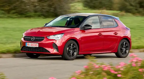 Roter Opel Corsa F fährt auf einer Straße, umgeben von grünen Feldern und blühenden Blumen. Keine Probleme mit diesem flotten Flitzer!