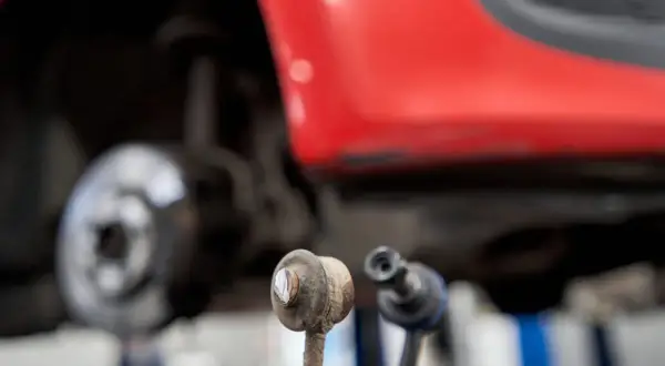 Zwei Koppelstangen liegen vor einem roten Auto. Eine Koppelstange ist defekt und muss ersetzt werden.