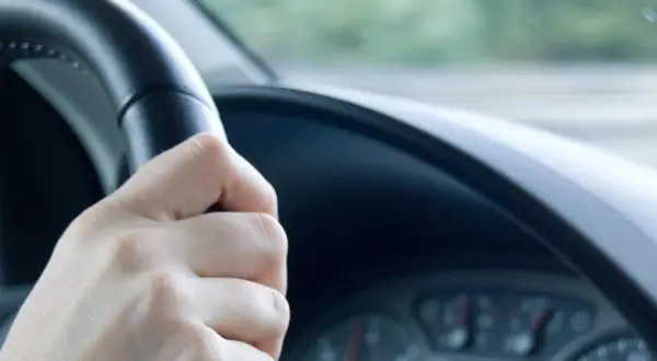 Hand am Lenkrad, das leicht flattert. Die Straße zieht vorbei, ein Gefühl von Bewegung.