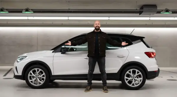 Mann neben weißem Seat Arona in Tiefgarage, lächelnd mit ausgestreckten Armen. Keine Seat Arona Probleme hier.