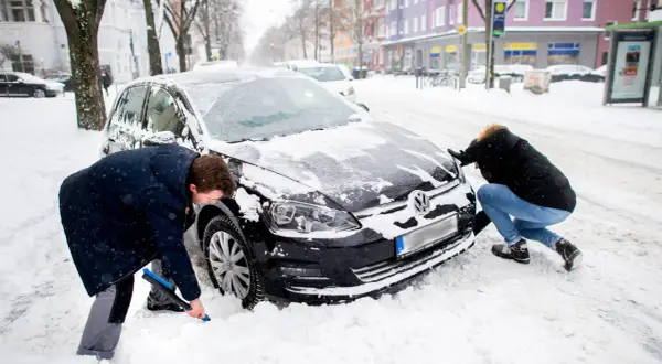 Zwei Personen schaufeln Schnee vom auto im winter. Ein Mann mit blauer Bürste reinigt das Rad, eine Frau schiebt die Motorhaube.