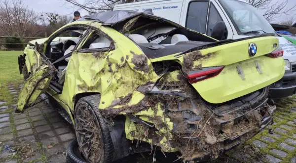 Ein gelber BMW mit Totalschaden, stark beschädigt und mit Schlamm bedeckt, steht neben einem weißen Lieferwagen.