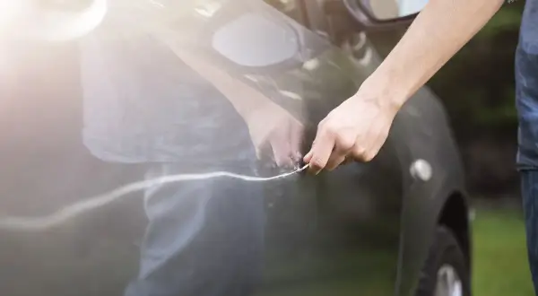 Hand mit Schlüssel verursacht Mikrokratzer am Auto. Die Sonne reflektiert auf dem dunklen Lack.