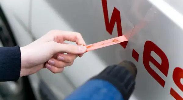 Hand zieht roten Aufkleber vom Auto ab, während ein Heißluftföhn das Entfernen erleichtert.