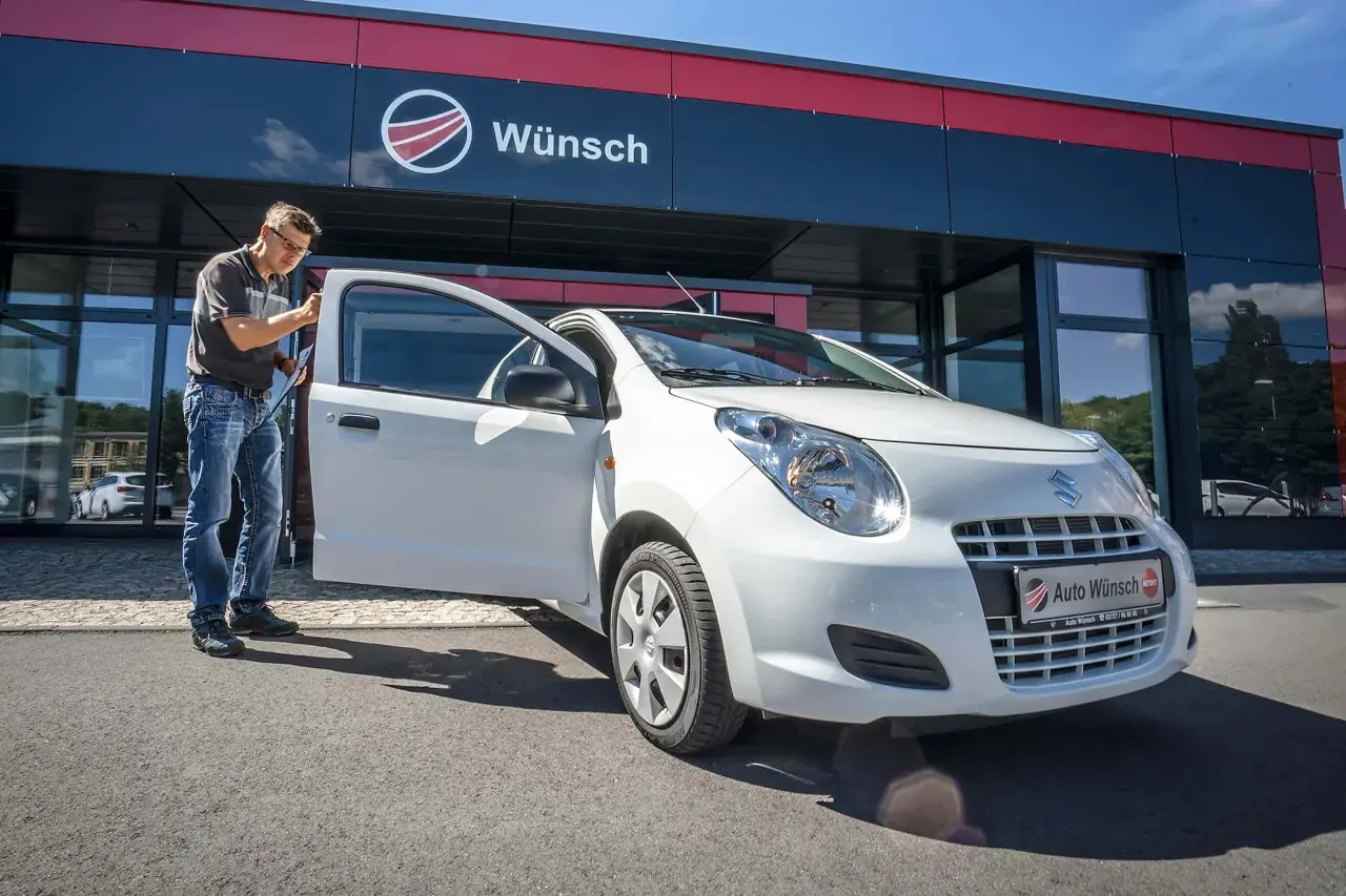 Mann prüft einen weißen Suzuki Alto vor dem Autohaus Wunsch. Mögliche Suzuki Alto Probleme werden hier behoben.